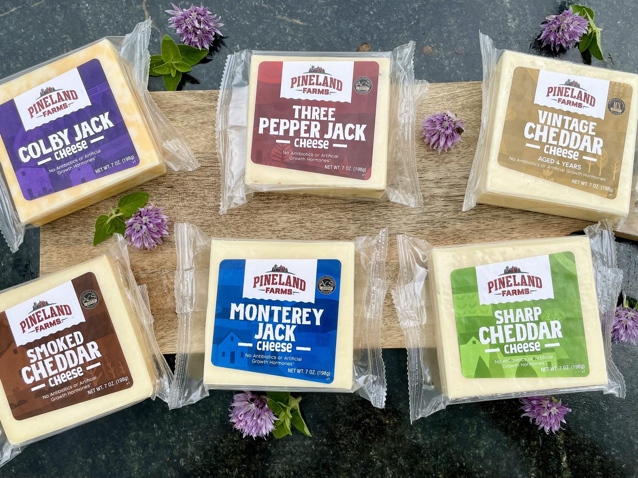 Multiple blocks of cheese on a butcher block table with scattered grapes, and fruit slices and a Pineland Farms Dairy sticker.